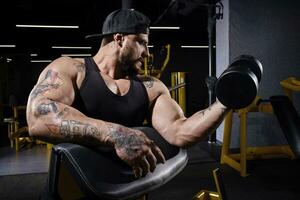 Tattooed guy with strong body, in black vest and cap. He lifting dumbbell, training his biceps, sitting on preacher curl bench at dark gym. Close up photo