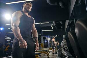 Tattooed, muscular bearded guy in black shorts, vest, cap. Looking at you, standing near set of black dumbbells. Dark gym. Sport. Close up photo