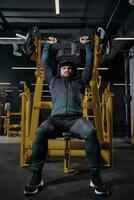 Male in black tracksuit with a hood, cap and sneakers. He performing chest press, sitting on an exercise machine, posing in dark gym. Full length photo