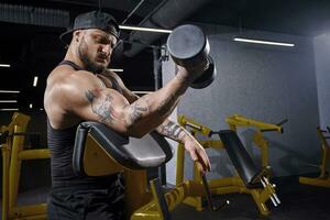 Bearded athlete in black vest and cap. He is lifting a dumbbell, training his biceps, sitting on preacher curl bench at dark gym. Close up photo