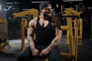 Tattooed, bearded athlete man in black sweatpants, vest and cap. He is looking thoughtful while sitting on an exercise machine at dark gym. Close up photo