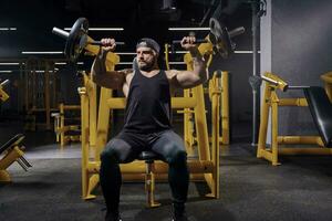 Tattooed, bearded sportsman in black sweatpants, vest and cap. He performing a chest press while sitting on an exercise machine at dark gym. Close up photo