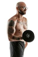 Athletic bald, tattooed man in black shorts is posing with a dumbbell isolated on white background. Close-up portrait. photo