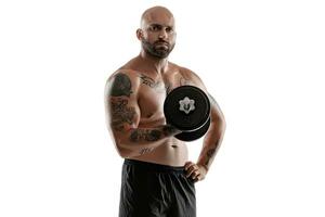 Athletic bald, tattooed man in black shorts is posing with a dumbbell isolated on white background. Close-up portrait. photo