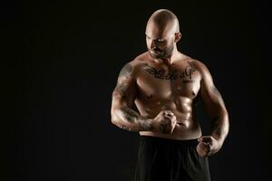 Athletic bald, bearded, tattooed man in black shorts is posing against a black background. Close-up portrait. photo