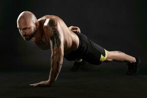 Athletic bald, tattooed man in black shorts and sneakers is posing against a black background. Close-up portrait. photo