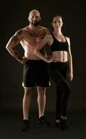 Athletic man in shorts and sneakers with brunette woman in leggings and top posing on black background. Fitness couple, gym concept. photo