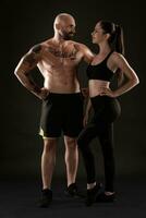 Athletic man in shorts and sneakers with brunette woman in leggings and top posing on black background. Fitness couple, gym concept. photo