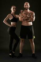 Athletic man in shorts and sneakers with brunette woman in leggings and top posing on black background. Fitness couple, gym concept. photo