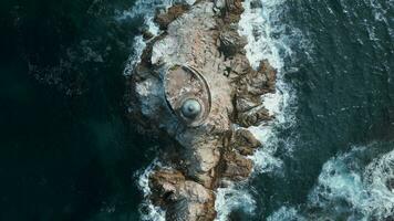 superiore Visualizza di faro su mare pietra. clip. abbandonato faro su bordo di roccioso promontorio nel mare. blu onde battere contro pietra ripiano con faro su soleggiato giorno video