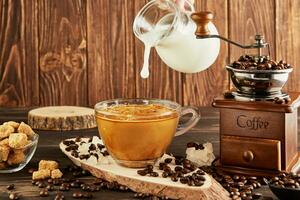 Pouring coffee from glass jug milk into glass cup with coffee, vintage coffee grinder and lump sugar on wooden background photo