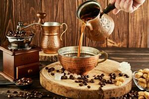 Pouring coffee from coffee maker into copper cup, an antique coffee grinder and copper milk jug on wooden background photo