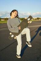 gracioso asiático niña disfrutando Patinaje, participación patineta me gusta guitarra y sombra jugando, teniendo divertido al aire libre foto