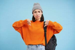 Upset asian woman shows thumbs down, dislike something, looking frustrated and disappointed, standing over blue studio background photo