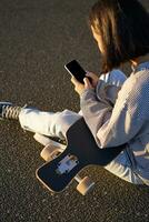 Vertical shot of asian girl typing, sending message or using mobile phone app, sitting on skateboard on road photo