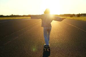 Carefree teen skater girl riding her skateboard with hands spread sideways, skating alone towards the sun photo