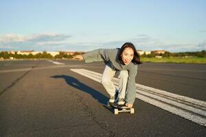 despreocupado patinador niña en su patineta, montando longboard en un vacío camino, participación manos oblicuo y riendo foto