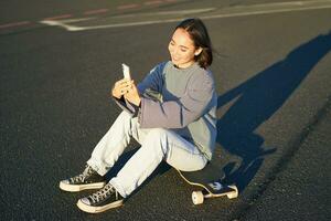 Happy asian girl sits on skateboard, takes selfie with longboard, makes cute faces, sunny day outdoors photo