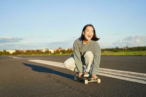 libertad y felicidad. contento asiático niña montando su longboard en un vacío soleado camino, riendo y sonriente, patinar foto