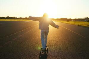 Rear view photo of young girl riding skateboard towards sunlight. Happy young woman on her cruiser, skating on longboard