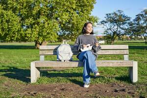 despreocupado niña se sienta en banco en parque con ukelele, obras de teatro y canta al aire libre en soleado contento día foto