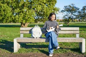 despreocupado niña se sienta en banco en parque con ukelele, obras de teatro y canta al aire libre en soleado contento día foto