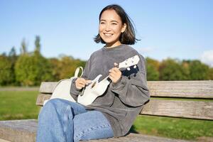 contento linda niña se sienta solo en banco en parque, obras de teatro ukelele guitarra y disfruta soleado día al aire libre foto
