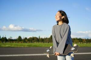 Portrait of asian woman with longboard. Korean girl skating, holding skateboard in hands, posing on road, smiling and looking aside photo