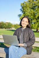joven asiático mujer trabajando de forma remota, Lanza libre niña se sienta en parque con computadora portátil, haciendo su trabajo desde al aire libre foto