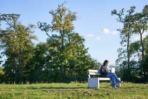 Side view of young woman sitting alone in park on bench, using her laptop to study or work remotely from outdoors photo