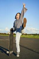 Excited asian girl dancing, standing with skateboard, skating on longboard and shouting carefree photo