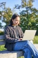 joven asiático mujer trabajando de forma remota, Lanza libre niña se sienta en parque con computadora portátil, haciendo su trabajo desde al aire libre foto