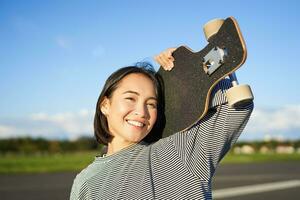 Happy and free asian girl holding cruiser board on shoulders and walking towards camera on empty road, skating on longboard and enjoying sunny weather photo