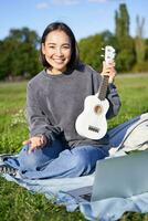 Vertical shot of happy asian girl plays instrument, shows her ukulele at laptop camera, video chats about music, teaches how to play, sits in park outdoors photo