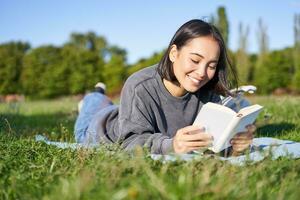 retrato de hermosa sonriente asiático chica, leyendo en parque, acostado en césped con favorito libro. ocio y personas concepto foto
