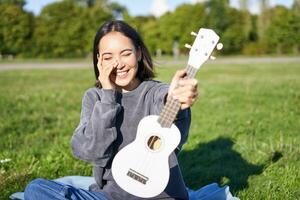 hermosa asiático niña con contento sonrisa, muestra su ukelele, se sienta fuera de en parque en césped, relaja con música foto