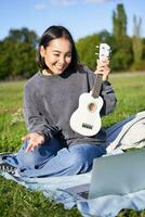 Vertical shot of happy asian girl plays instrument, shows her ukulele at laptop camera, video chats about music, teaches how to play, sits in park outdoors photo