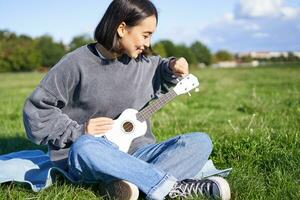 Singing asian girl playing ukulele on grass, sitting on blanket in park, relaxing outdoors on sunny day photo