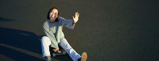 Positive korean girl covers her face from sunlight, sits on skateboard and smiles happily photo