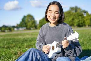 despreocupado asiático niña canto y jugando ukelele en parque, sentado en césped, músico relajante en su gratis hora al aire libre foto