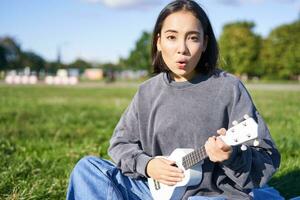 Portrait of cute asian girl with musical instrument. Young woman with surprised face, holding ukulele and sitting in park on blanket photo