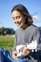 vertical Disparo de sonriente asiático niña canto y reír, jugando ukelele, aprender cómo a jugar instrumento, sentado al aire libre en parque foto