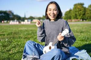 retrato de emocionado asiático chica, sentado con ukelele en parque en verde césped, mirando asombrado y sorprendido con bueno Noticias foto