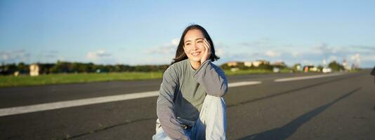 Vertical shot of asian girl skater, sits on her skateboard and smiles, enjoys sunny day, cruising on longboard on empty road outdoors photo