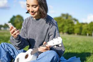 tecnología y música. sonriente asiático hipster niña sentado en parque con teléfono inteligente y participación ukelele, jugando instrumento foto