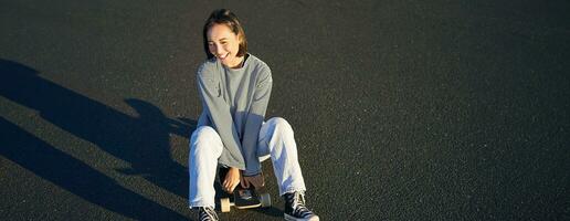 Portrait of beautiful asian girl skating, sitting on her skateboard and smiling. Cute teenager with longboard, skating on road photo