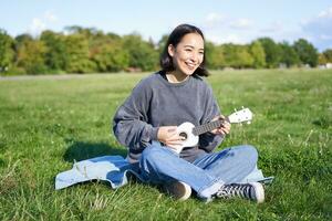 contento morena asiático chica, estudiante jugando ukelele sentado en césped en parque, relajante, canto canción, estilo de vida concepto foto