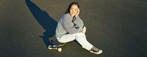 Portrait of asian woman sitting on skateboard, skating on her cruiser longboard, using smartphone app photo
