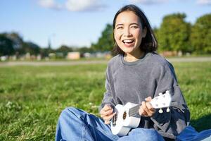 Portrait of asian girl student, playing ukulele and singing in park, sitting alone on blanket and enjoying making music photo