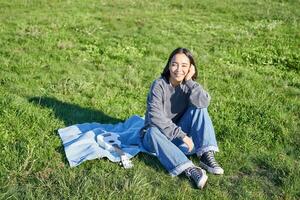joven asiático niña disfrutando soleado día al aire libre. contento estudiante teniendo picnic en césped en parque, jugando ukelele foto